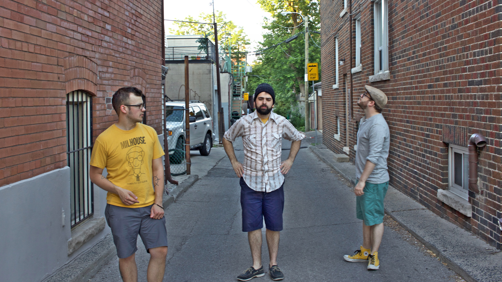 brick walls line a paved alley, with three young men in shorts and t-shirts standing at varied distances from the camera