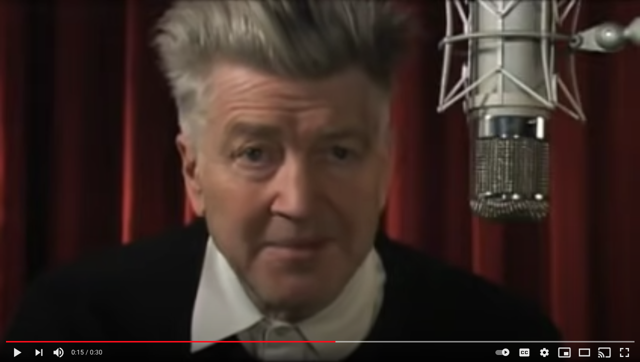 close up of director david lynch's face with a microphone in front of him and a red curtain in the background