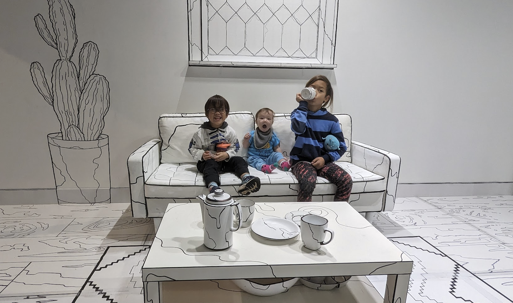 Two boys and one baby girl pose with different silly faces on a white couch with black outlines in a room designed to look like a line illustration on pape