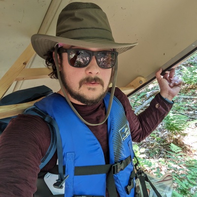 Bearded white man with an adventure hat, sunglasses, a life jacket, and a canoe on his shoulders